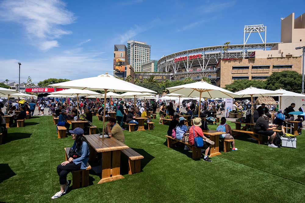 Petco Park in the background with benches with umbrellas, people sitting on the benches, on top of astroturf at the Petco Interactive Zone