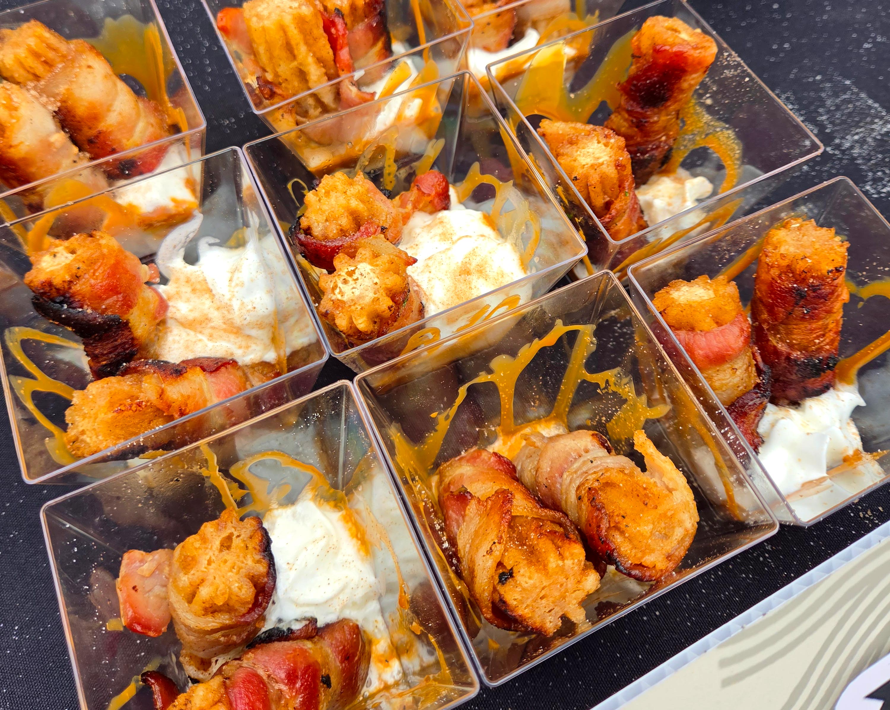 Multiple clear containers are filled with bacon wrapped churros and a side of sauce on a table.