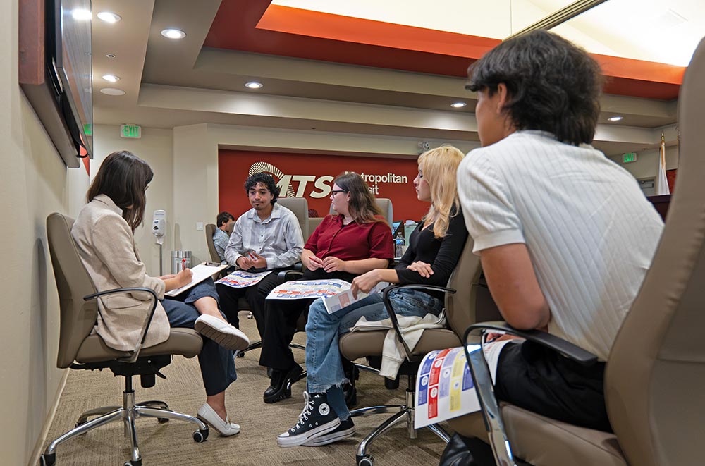 5 people are sitting together in a semicircle. One is writing and the other four are facing her, holding a paper with graphics on it. They are in the MTS board room.