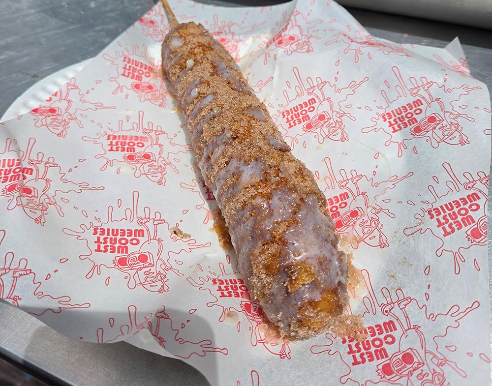 A long churro dog covered in sauce and cinnamon on a piece of white paper that has West Coast Weenies logo all over the paper.