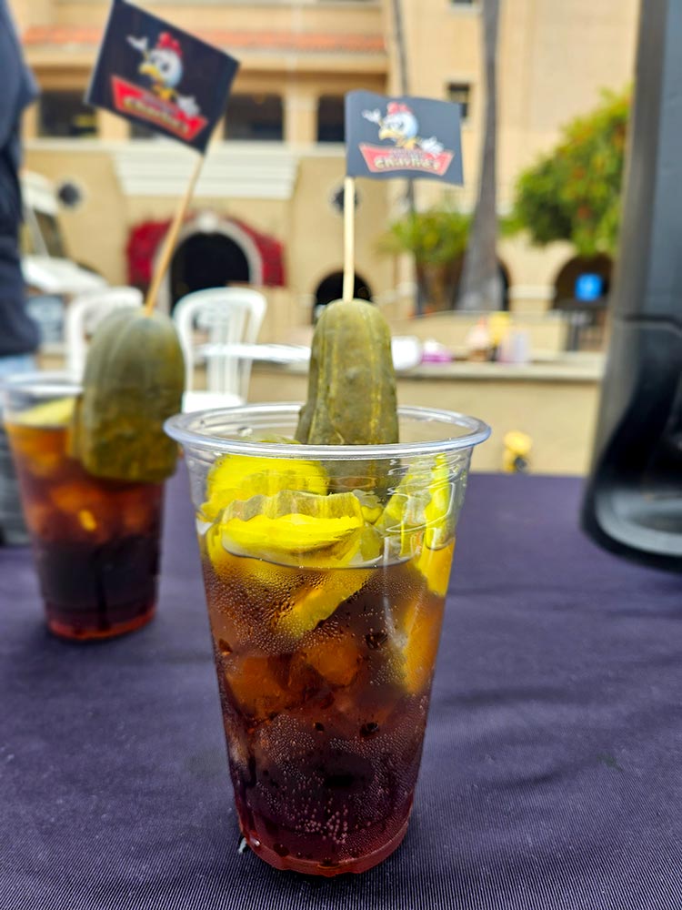Plastic cup filled with Dr Pepper soda, pickle slices, and a whole pickle.