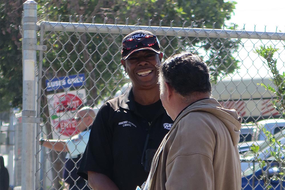 A woman in uniform smiling and stands talking to an individual in front of a fence.