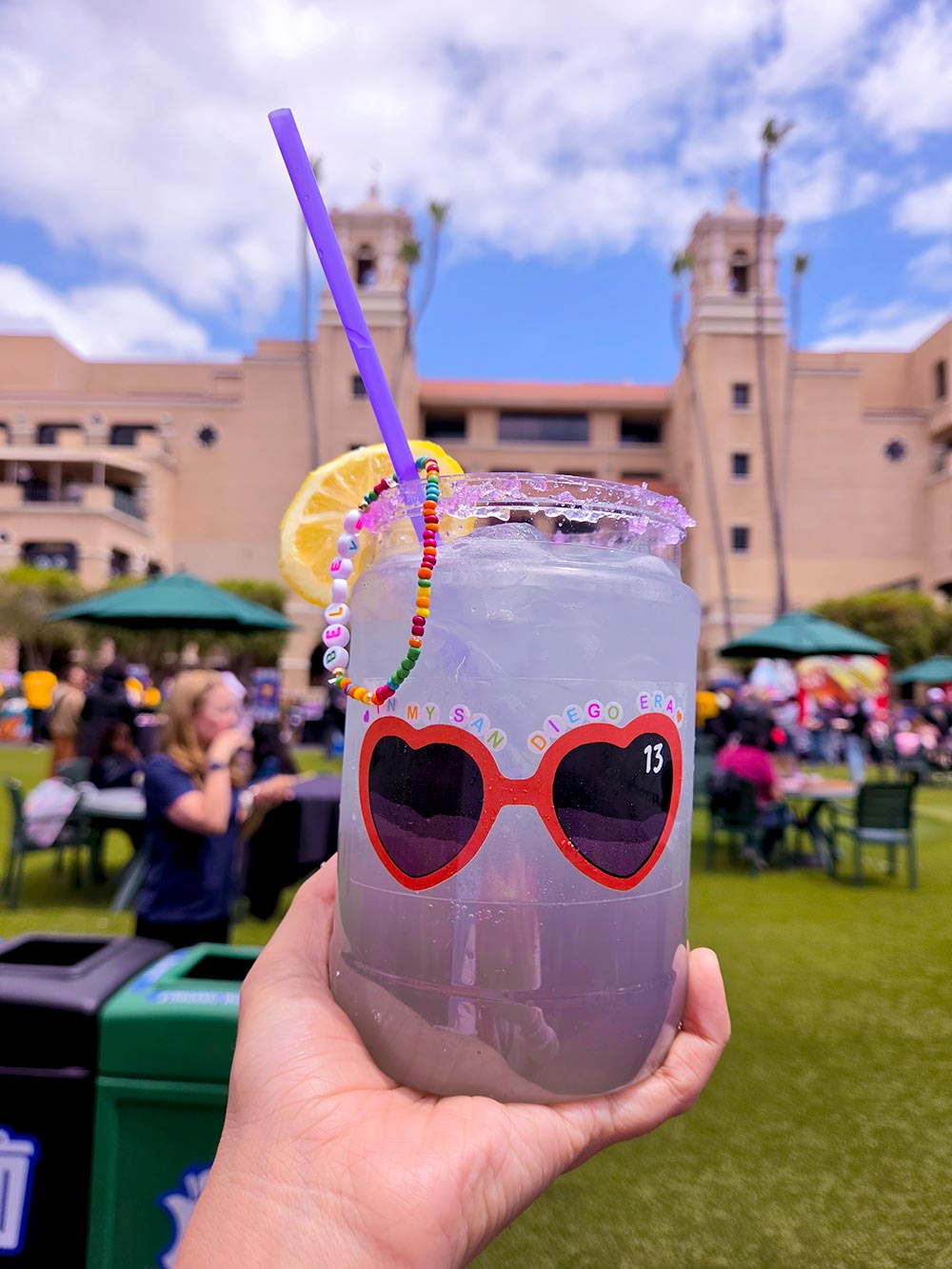A plastic jar of lemonade with a friendship bracelet dangling from the straw on a grass plaza.
