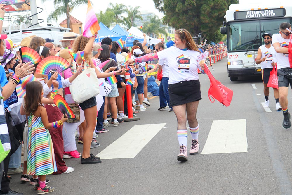 A woman in an MTS Pride Staff T-shirt hands out a giveaway during the Pride Parade as participants walk along the street, spectators watch from the sidewalk, and an MTS bus appears in the background.