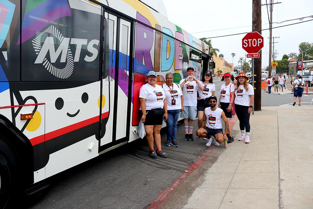 Seven MTS staff members wearing matching white MTS Pride t-shirts stand in a group next to the MTS Bus wrapped with Ride to Pride and the MTS mascot.