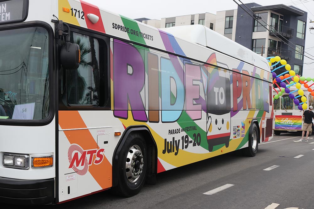 MTS bus with a wrap on the outside that says 'Ride to Pride Parade and Festival' with the bus mascot holding a Pride flag