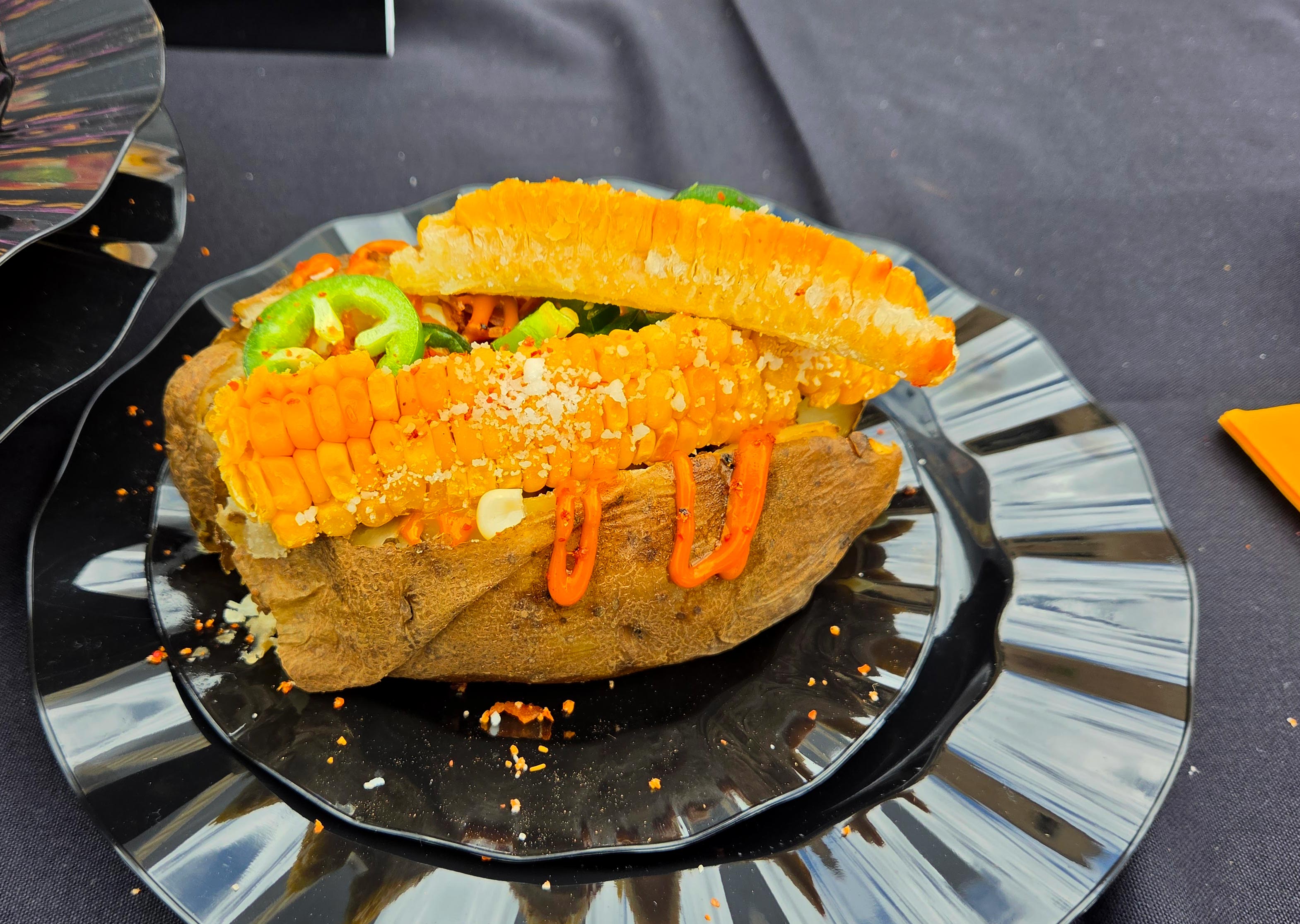 A black table with a black plate with a baked potato on it, with corn nibs and jalapenos showing on top.