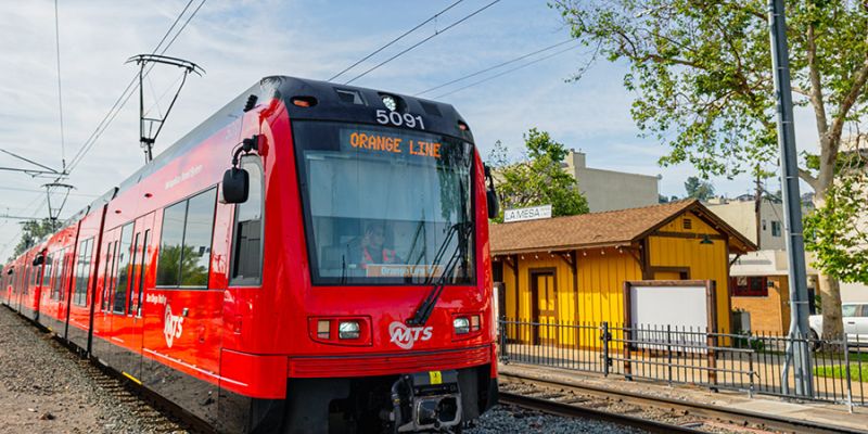 Trolley on the Orange Line in La Mesa on the tracks with a building and trees next to it