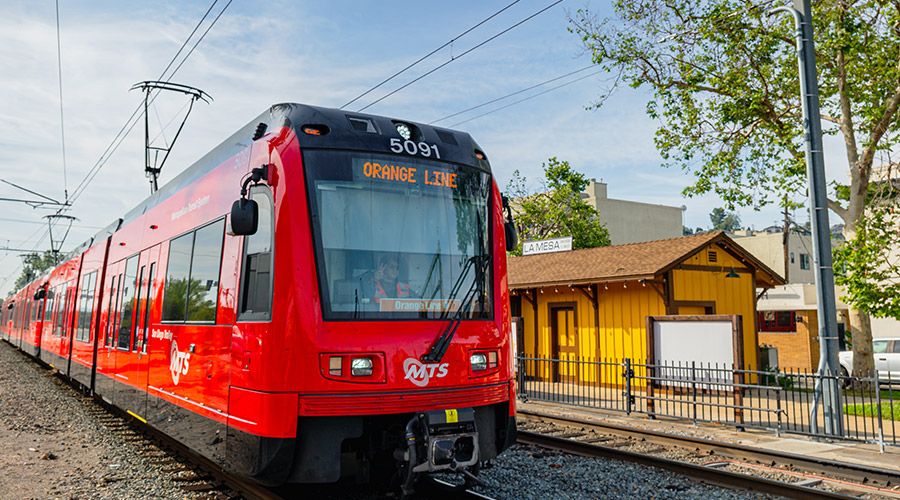 Trolley on the Orange Line in La Mesa on the tracks with a building and trees next to it