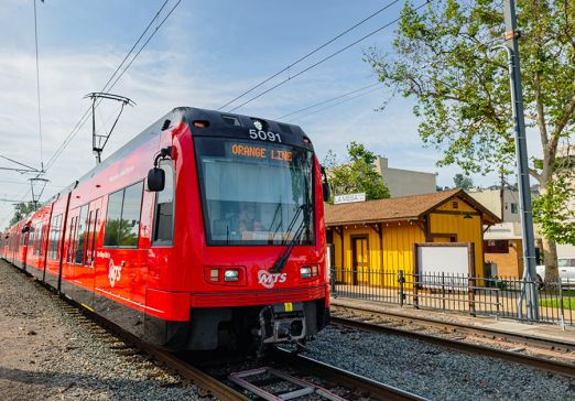 Trolley on the Orange Line in La Mesa on the tracks with a building and trees next to it