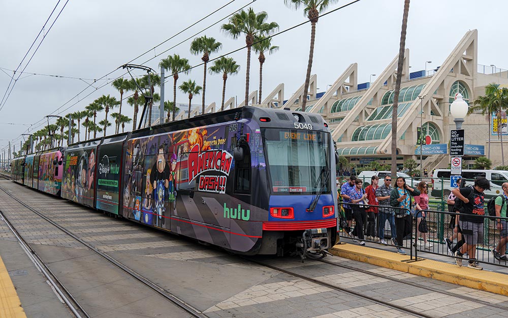 The Trolley stops at Convention Center Station with a crowd of people walking by. The San Diego Convention Center is in the background.