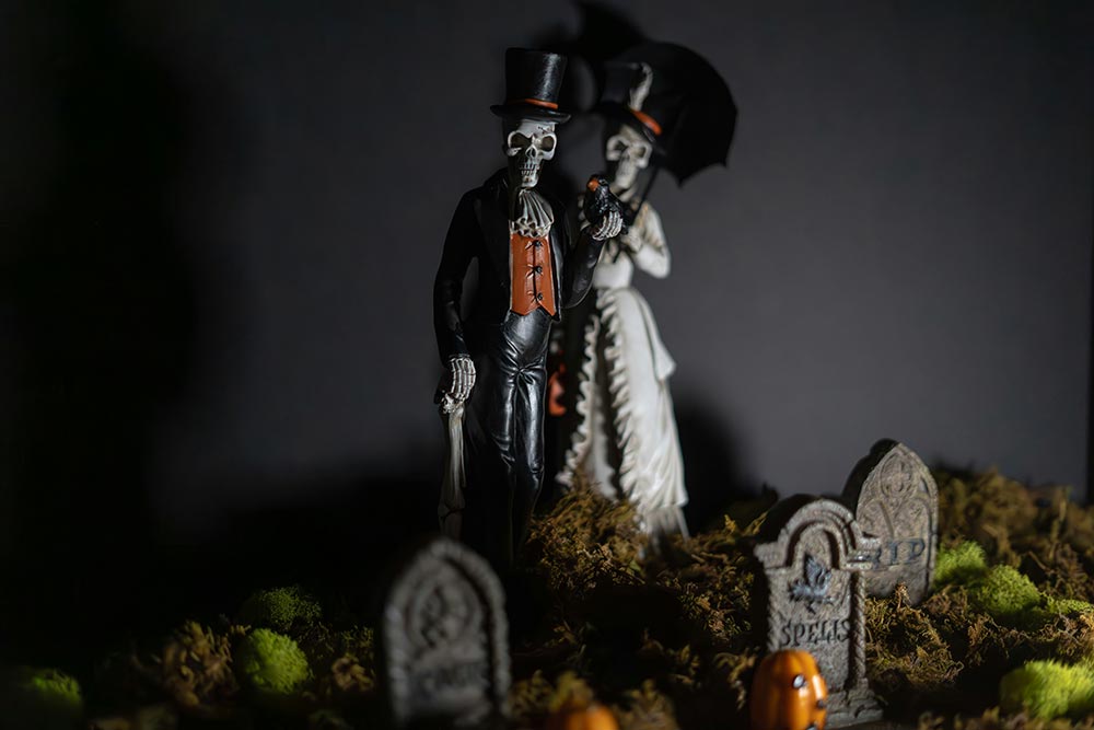 Two skeletons dressed up, one in a suit and one in a dress holding an umbrella, in a graveyard with tiny headstones
