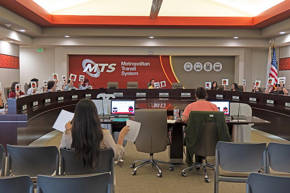 Inside the MTS board room. There is a round dias and people are seated around the dias, holding up two different images. In front is a woman on a laptop and a second woman also holding sheets of paper.